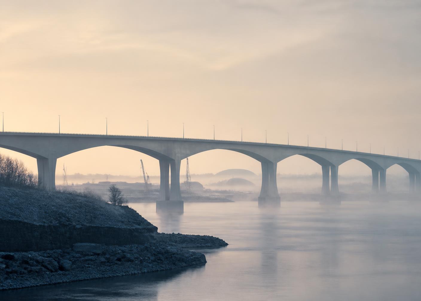 Ponte de concreto sobre rio em névoa, ao amanhecer, junto a um canteiro de obras